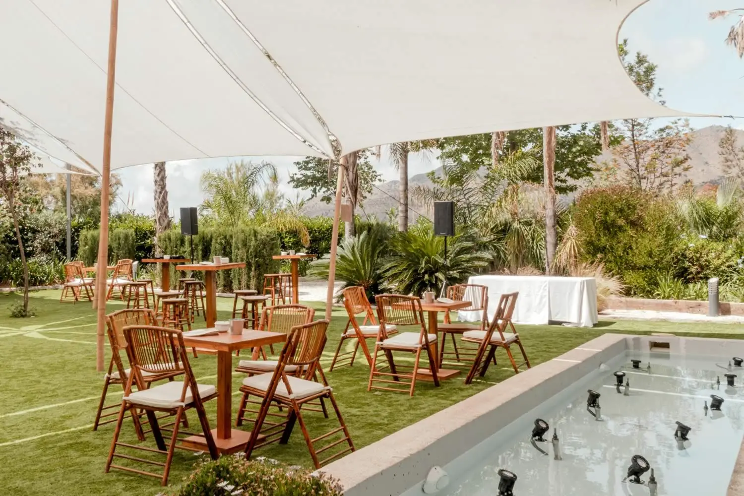 Terraza de cóctel bajo velas blancas, con mesas y sillas de madera junto a un canal de agua